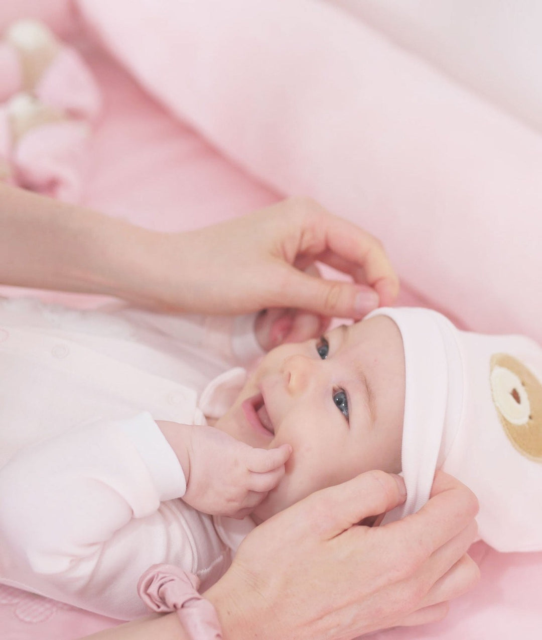 Adult hand adjusting white baby beanie with bear patch on smiling infant in onesie