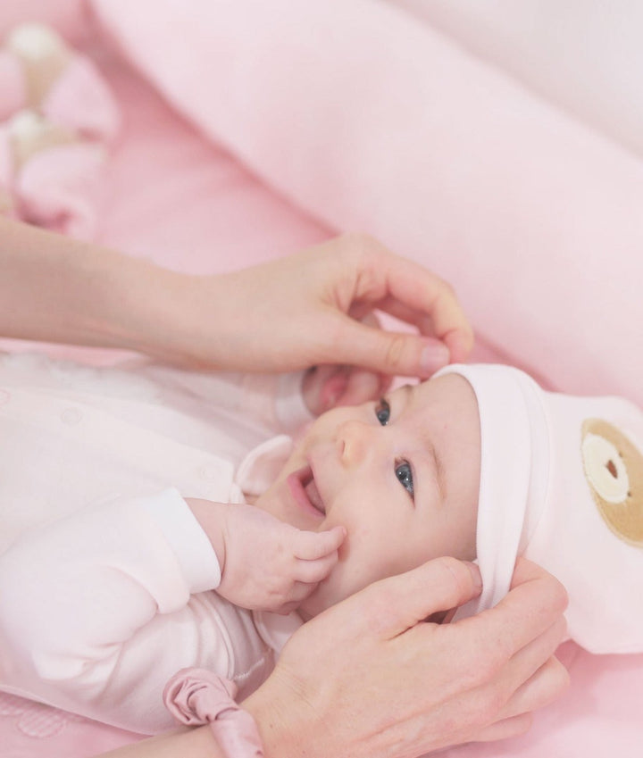 Adult hand adjusting white baby beanie with bear patch on smiling infant in onesie