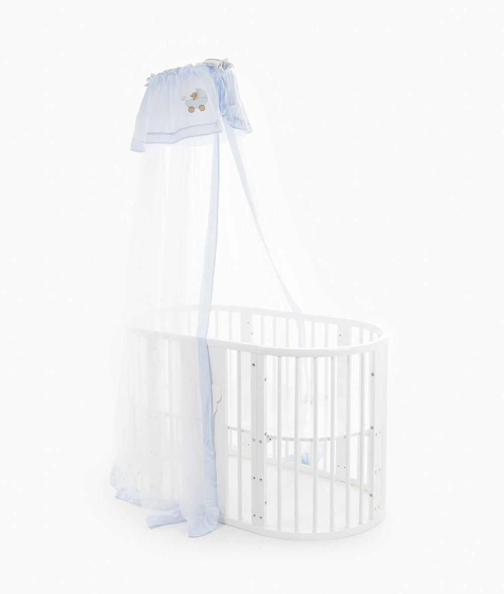 Side profile of translucent white netting cot canopy cascading from blue trimmed header to base of oval white crib