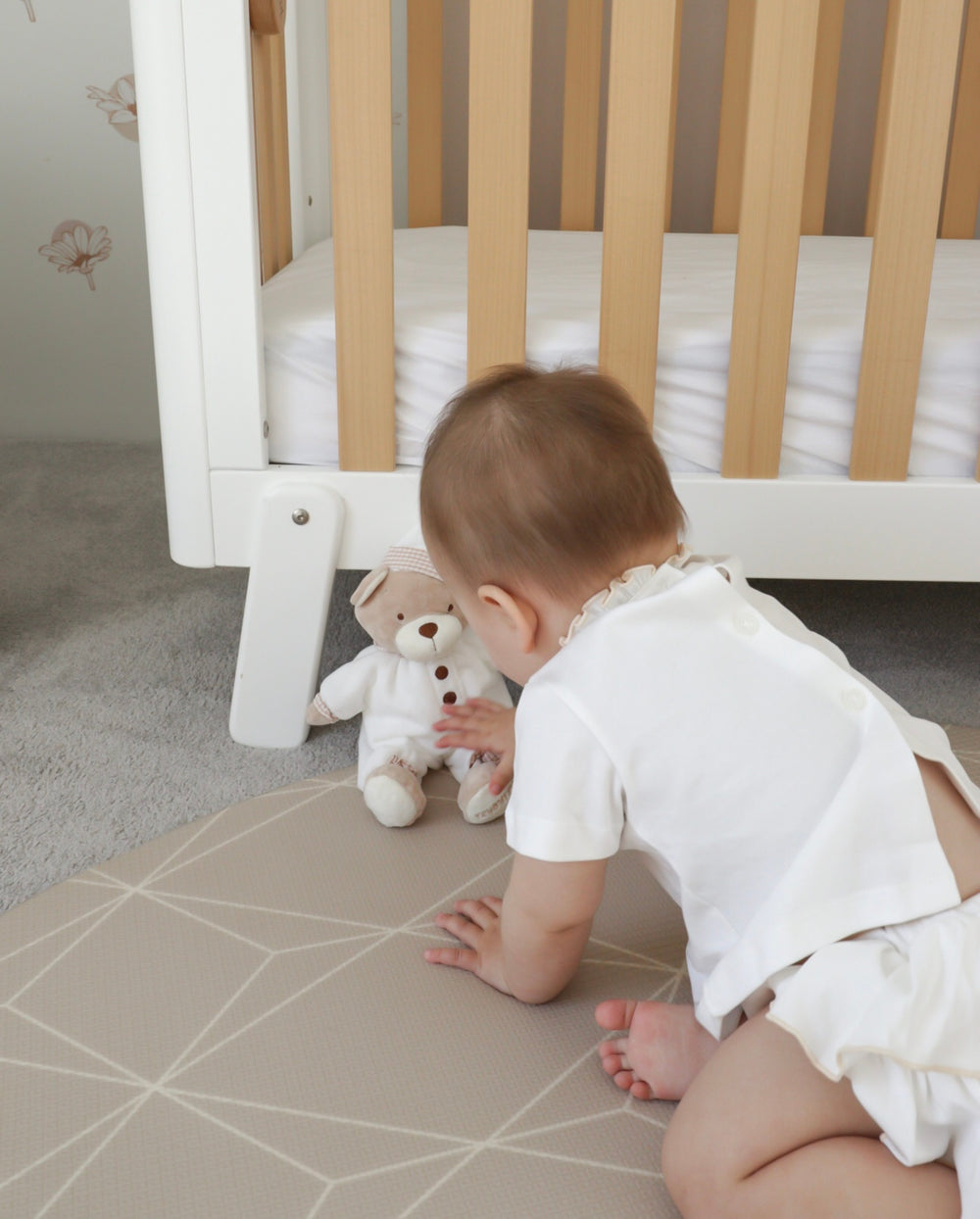 Baby crawling on geometric mat reaching for Cuddle Bear teddy in nightcap near crib