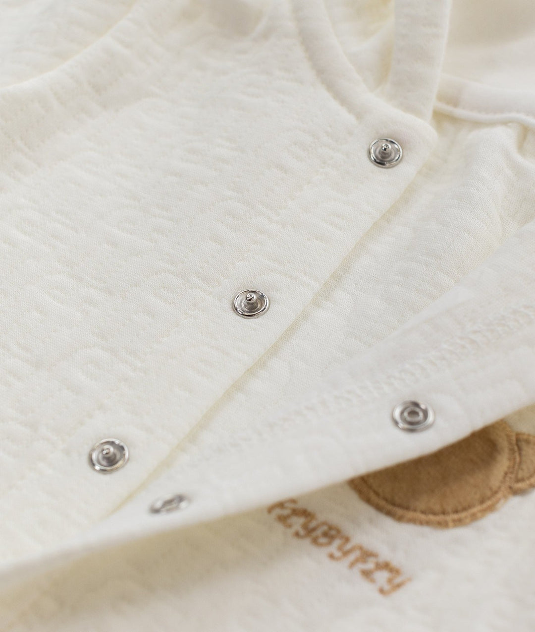 Close up of metallic silver snap buttons on off-white cotton jumpsuit placket next to tan bear embroidery and brand text