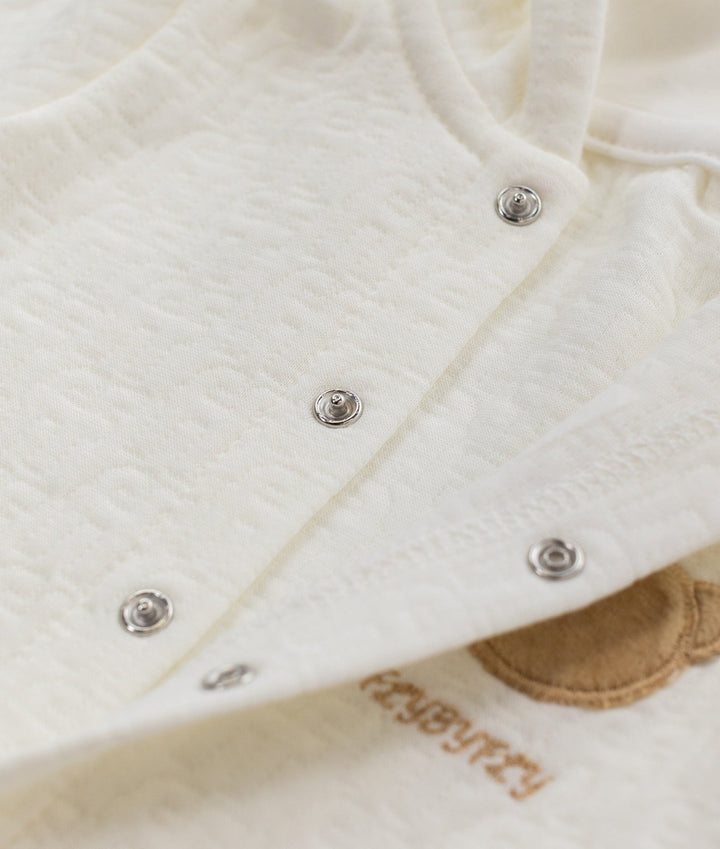 Close up of metallic silver snap buttons on off-white cotton jumpsuit placket next to tan bear embroidery and brand text