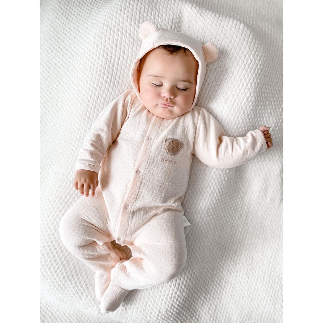 Infant wearing light pink hooded bear jumpsuit on white knitted blanket