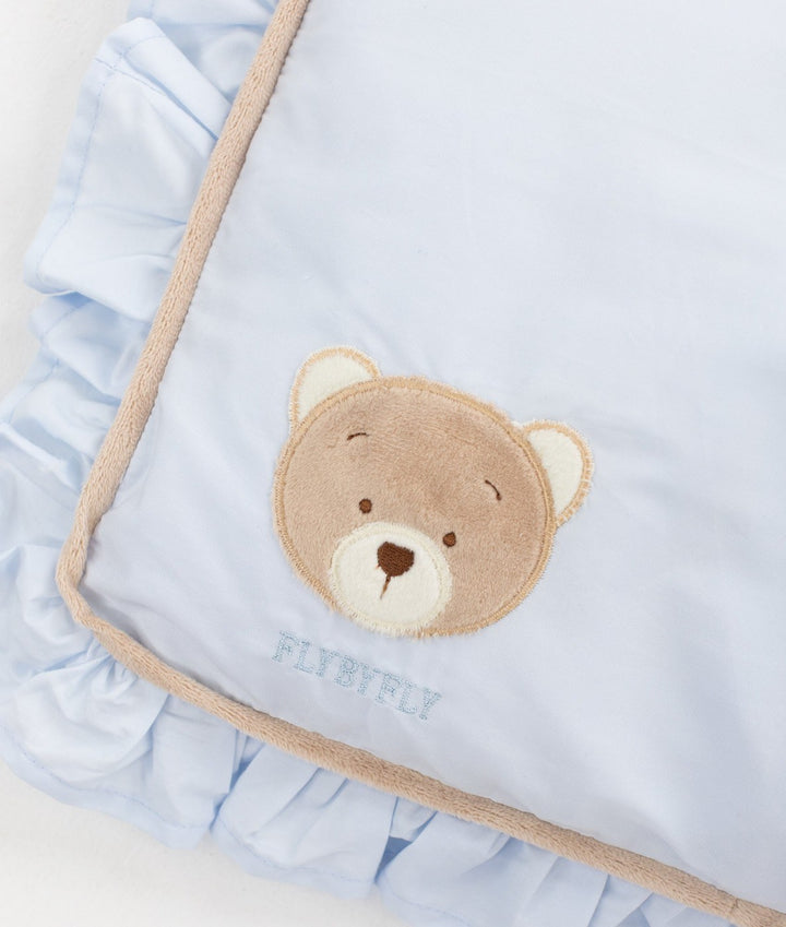 Pale blue bedding detail showing textured brown teddy bear face applique with cream ears, embroidered FLYBYFLY text, tan piping, and ruffled edge